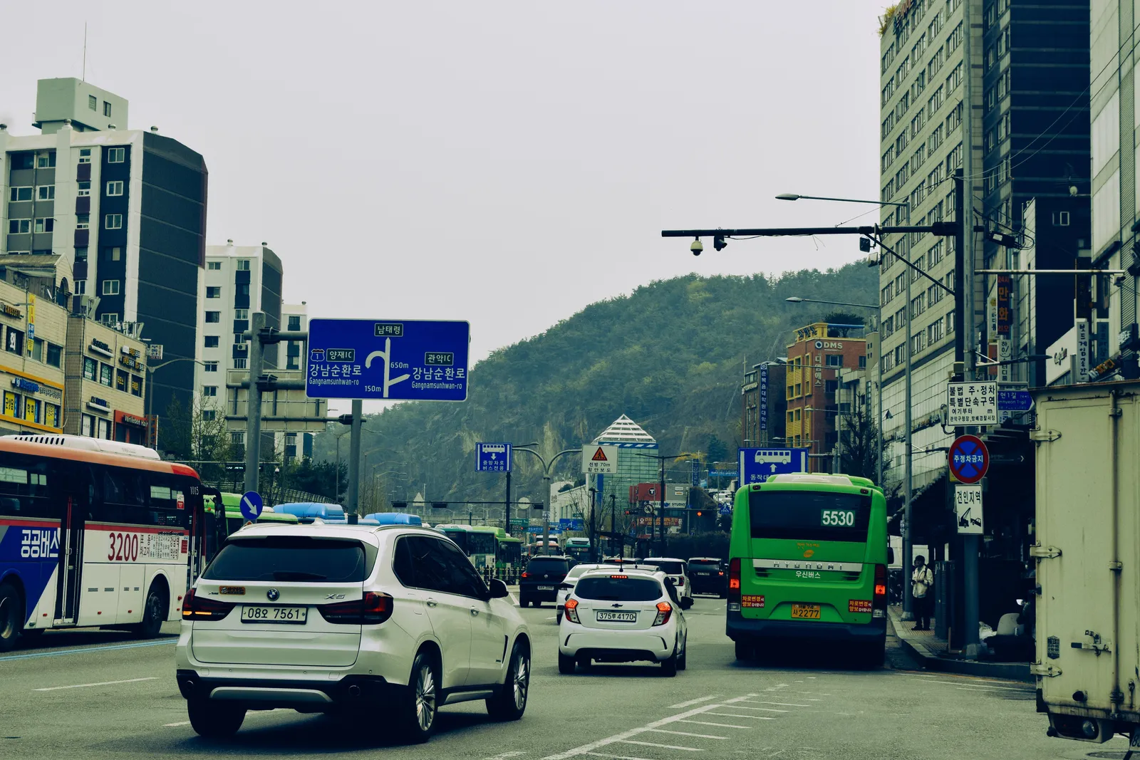 A yellow taxi and city buses at a busy street crossing beside a subway entrance in Seoul, South Korea