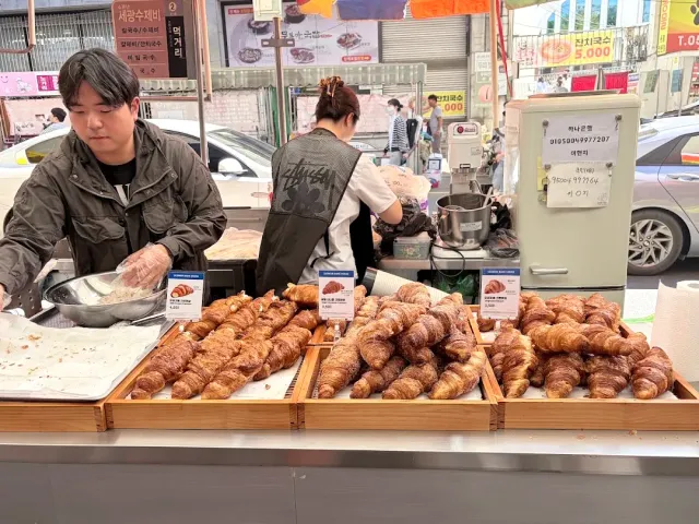 A market stall with wooden trays full of croissants, vendors behind it and Korean signs in the background