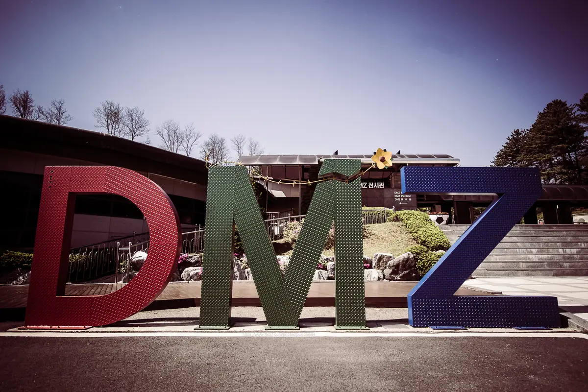 Giant coloured letters standing on a plaza in front of a low exhibition building and pine trees