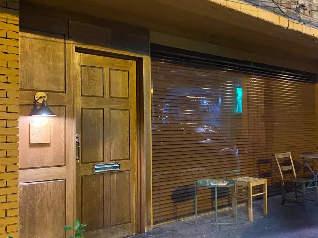 A wooden door lit by a wall lamp at night, beside a wooden blind and a small table