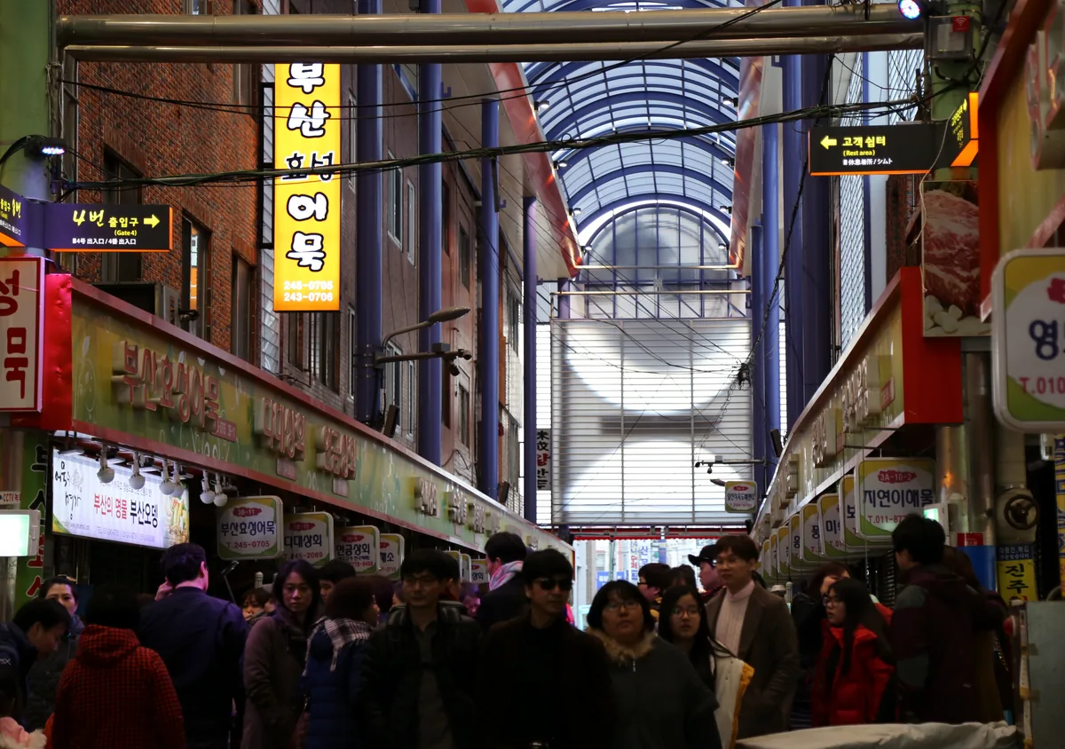 A covered market arcade in Busan with a vaulted glass roof, yellow eomuk stall signs and a crowd of shoppers