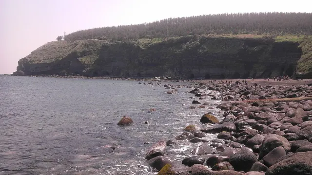A dark pebble shore facing a cliff with dark tunnel openings, greyish sea