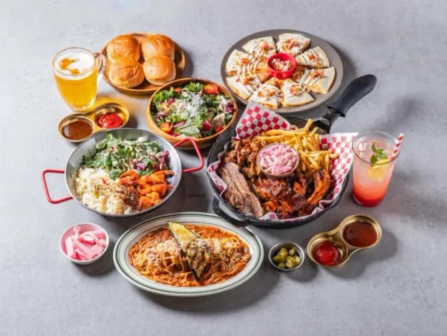 An assortment of dishes on a grey surface, among them smoked meat with fries, pasta, salad and a glass of beer
