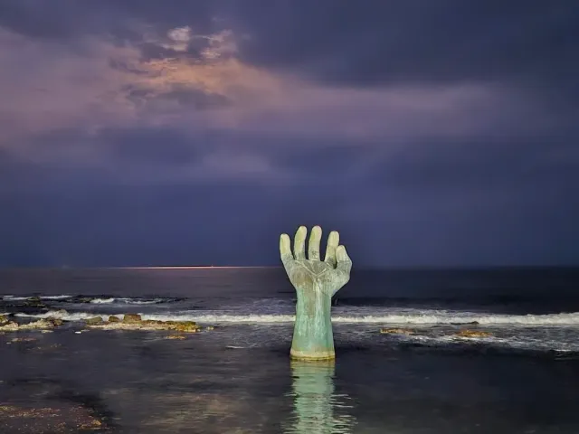 A giant hand sculpture rising out of the water, purple cloudy sky