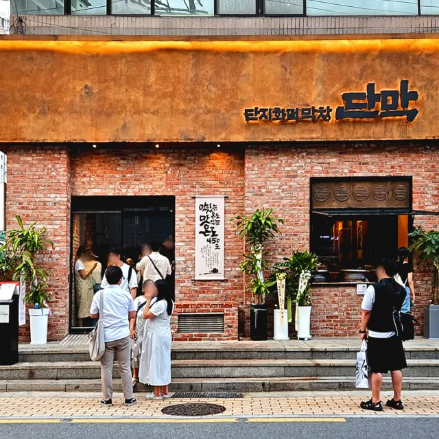 A brick front with a mustard coloured sign, people waiting at the entrance and planters on the steps