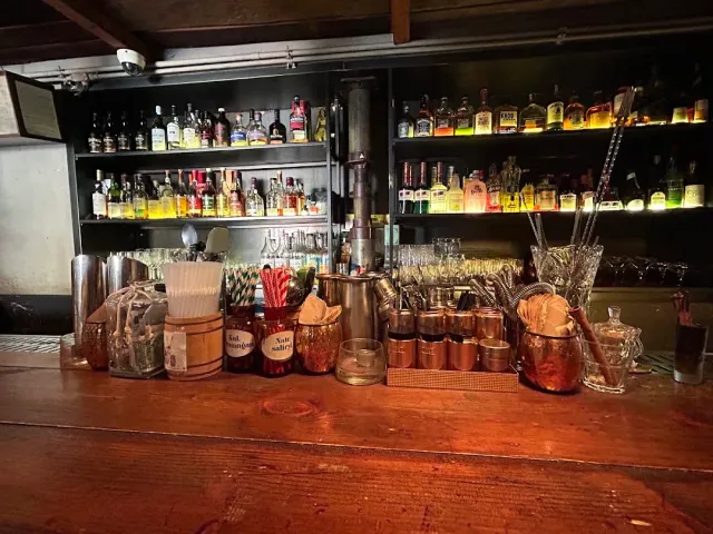 A counter with metal mugs, jars of straws and mixing tools, and lit bottle shelves behind