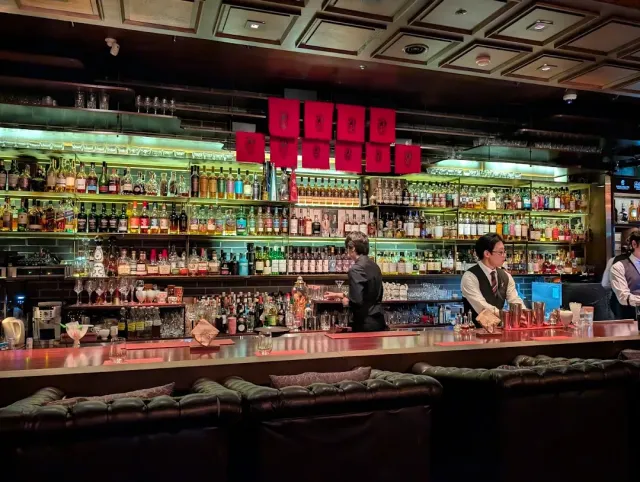 A long bar with bartenders, a wall of shelves loaded with bottles and leather sofas in front