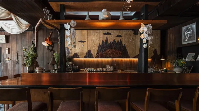 A long wooden bar counter with chairs, white orchids and a lit mountain painting behind