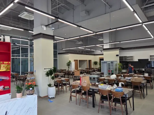A large hall with light strips on the ceiling, rows of tables and chairs and drinks fridges
