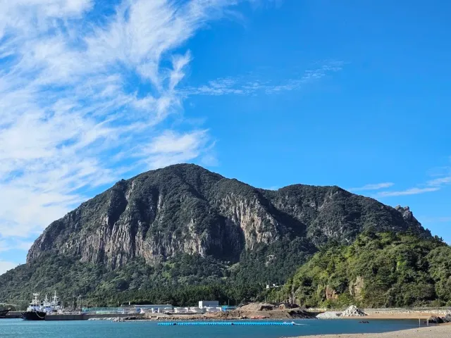 A forested rock mountain rising above a small harbor and a strip of shore, blue sky
