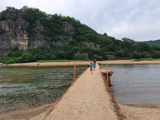 A narrow sand path crossing a river toward a forested cliff, walkers on it