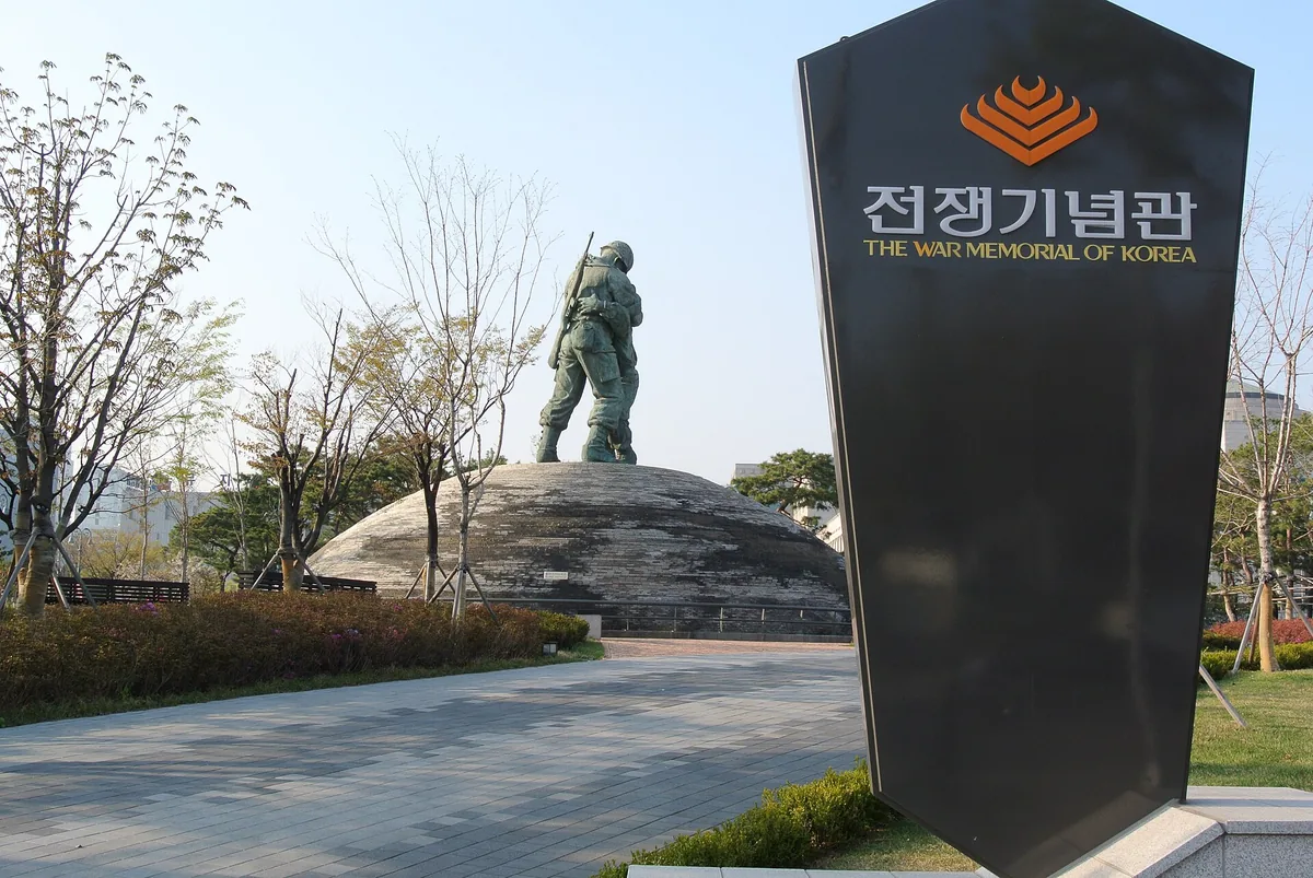 A dark name stone at a museum entrance, with a bronze statue of a soldier on a stone mound behind it