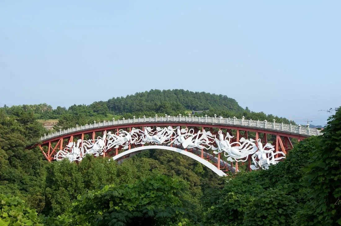 Red arched bridge lined with white figure sculptures spanning a green wooded gorge