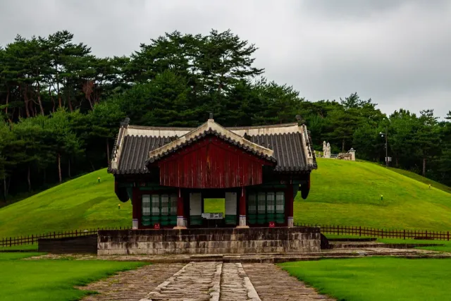 A red wooden structure with a tiled roof before a rounded grass mound and forest