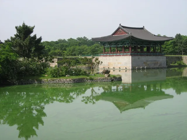 A traditional wooden pavilion reflected in a green pond ringed with trees