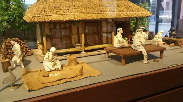 A display case model of a thatched roof house with figures in traditional white clothing in the yard