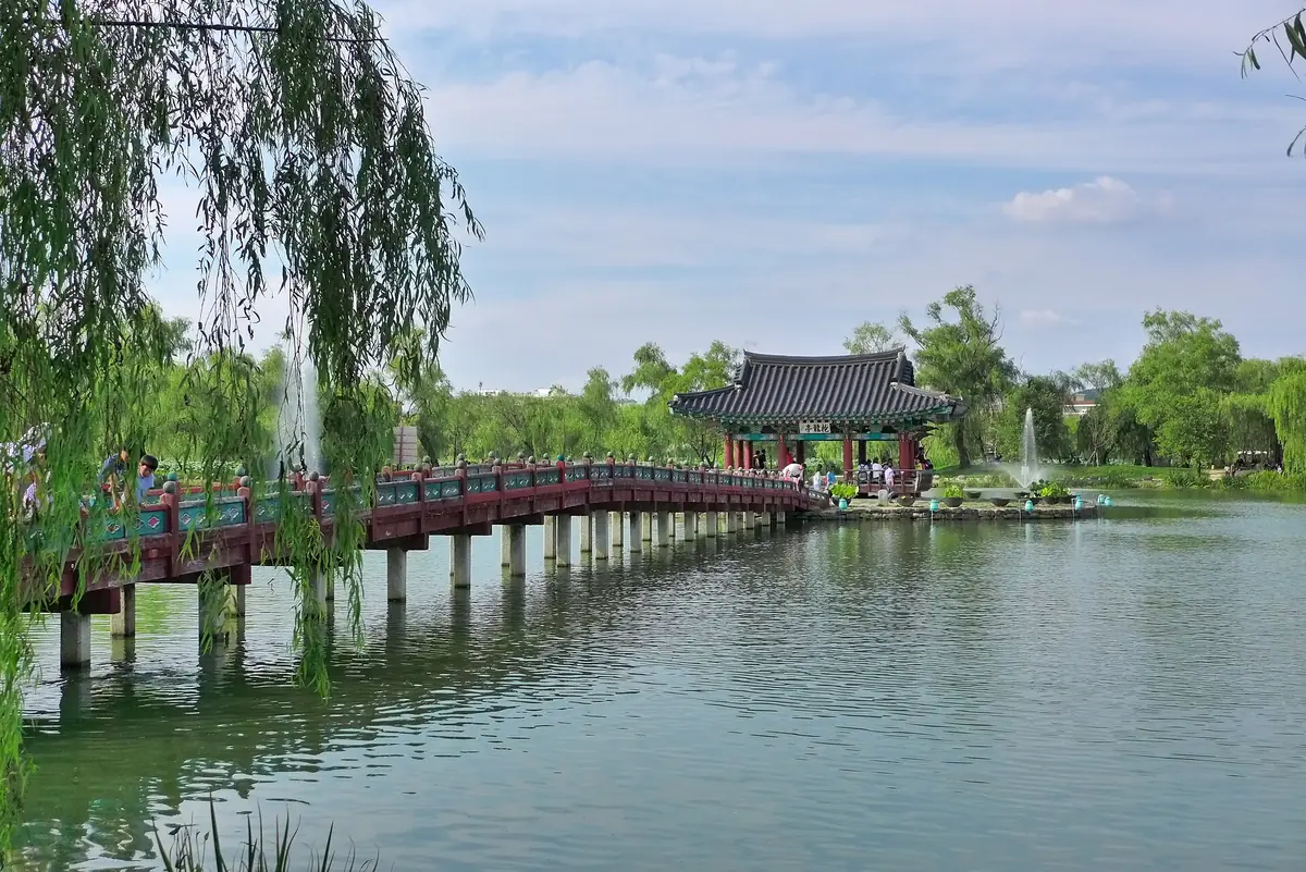Curved wooden bridge leading to a traditional pavilion on a lake, framed by weeping willow branches