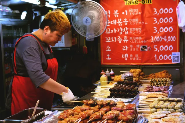 A vendor arranging skewers and tempura on a stall counter, an orange price sign