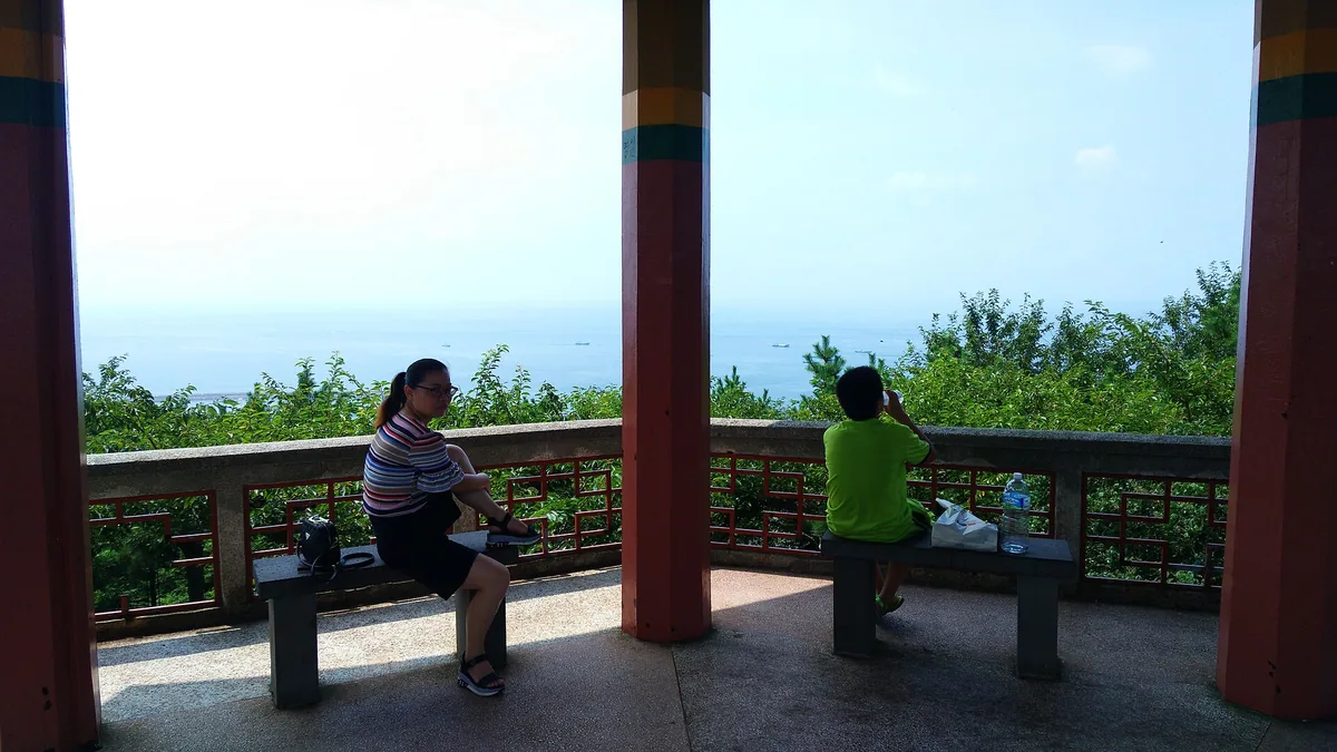 Two visitors leaning on a railing above the sea on Jeju, beside a red timber post