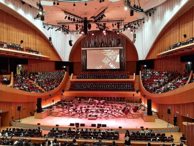 A concert hall full of audience, an orchestra on the stage and curved wooden walls