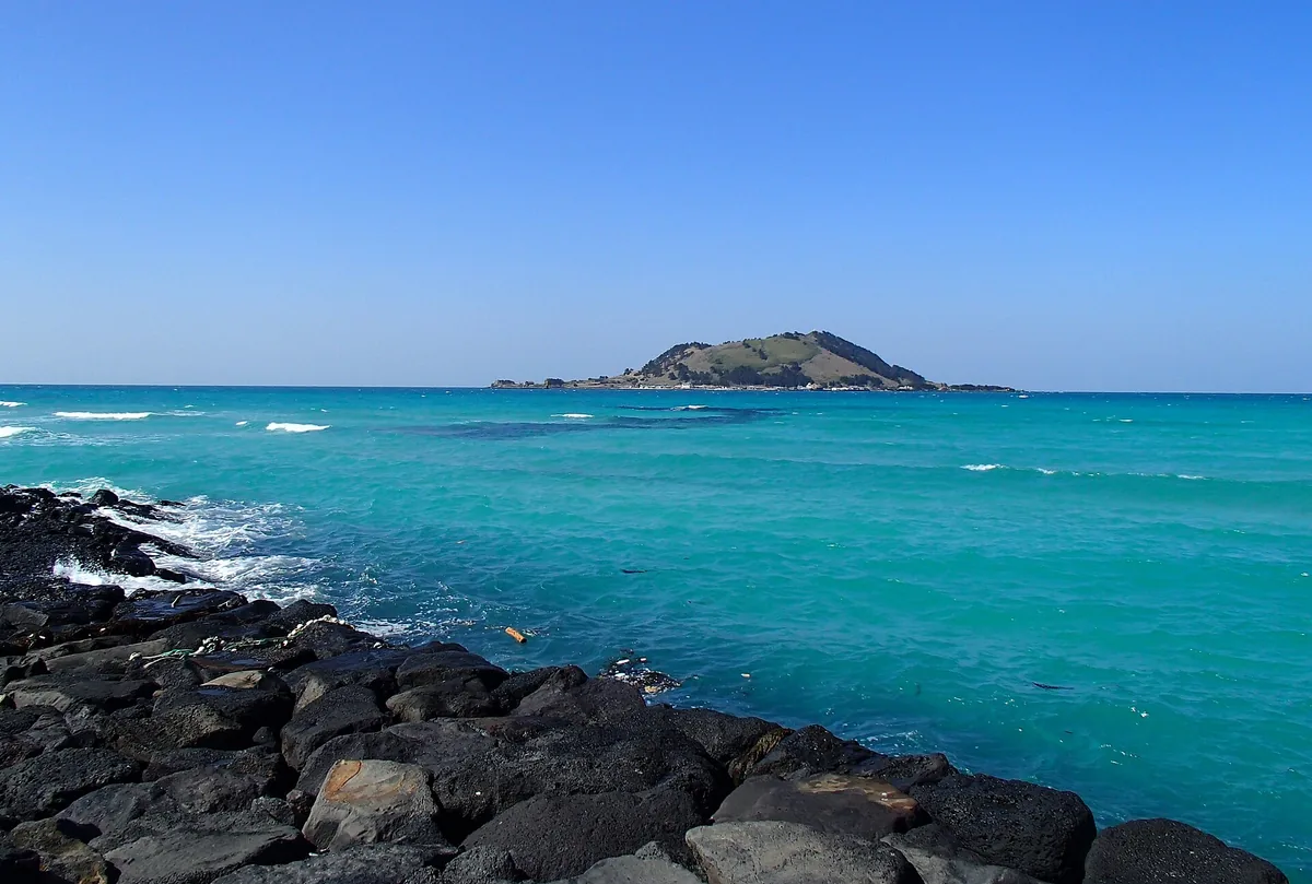 Turquoise water beside black volcanic rocks, with a small green island on the horizon