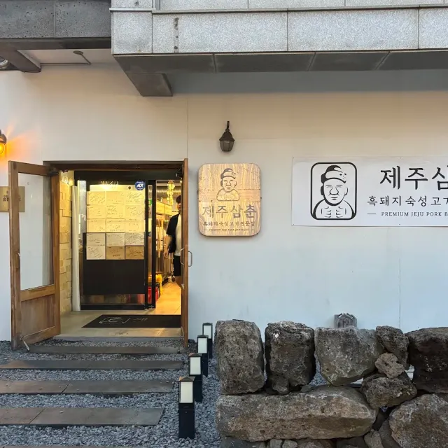An entrance in a white facade with an illustrated wooden sign, an open door and a basalt stone wall