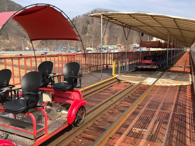 A red pedal cart with a canopy on the rails, and a long shed in the background