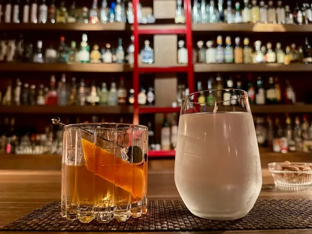 Two cocktail glasses on a counter, with lit bottle shelves and a red ladder behind them
