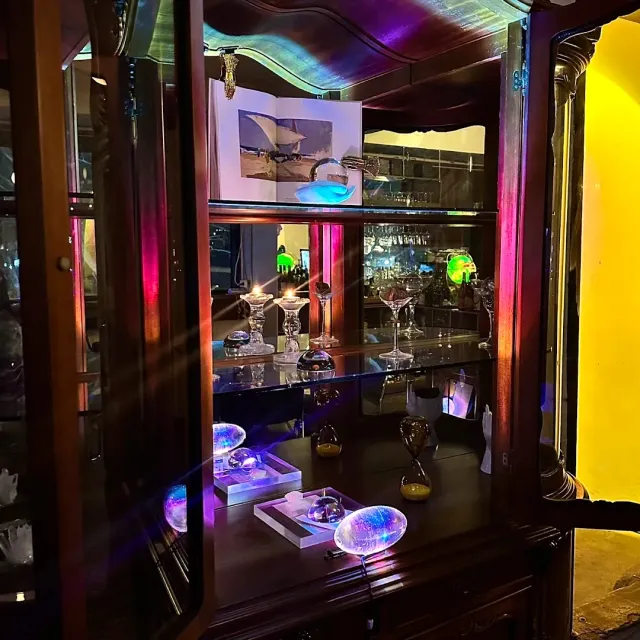 A dark wooden cabinet under coloured light holding shimmering glass pieces and cocktail glasses
