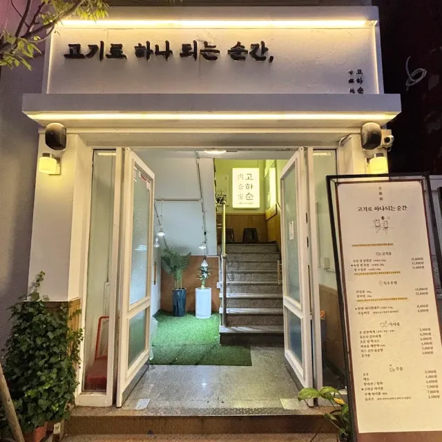 A lit entrance in the evening with a sign, open doors, stairs inside and a menu board