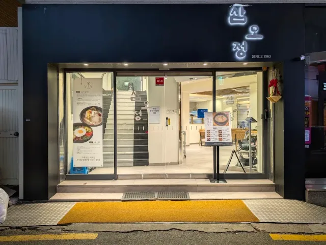 A dark facade with a lit sign, open glass doors and a lit staircase inside