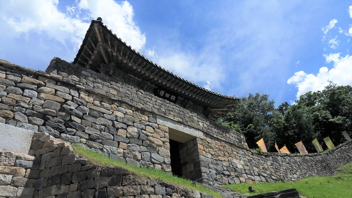 Stone gateway in a fortress wall topped by a timber pavilion against a blue and cloudy sky