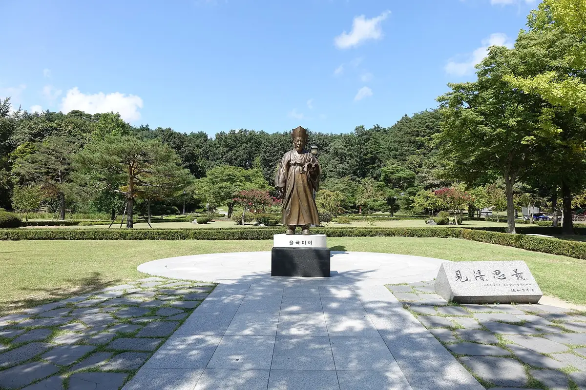 A bronze statue of the scholar Yulgok Yi I on a stone plinth on a clipped lawn, wooded hills behind