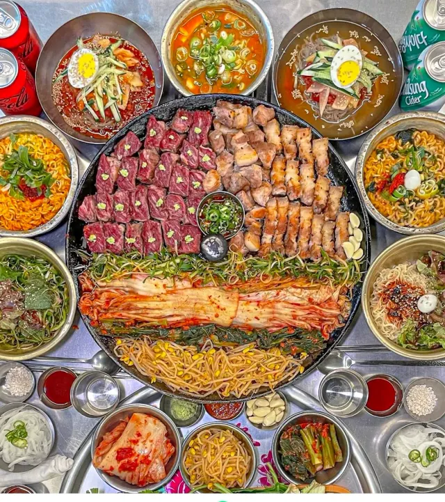 A top view of a round grill tray with cuts of meat, spicy cabbage and bowls of side dishes around it