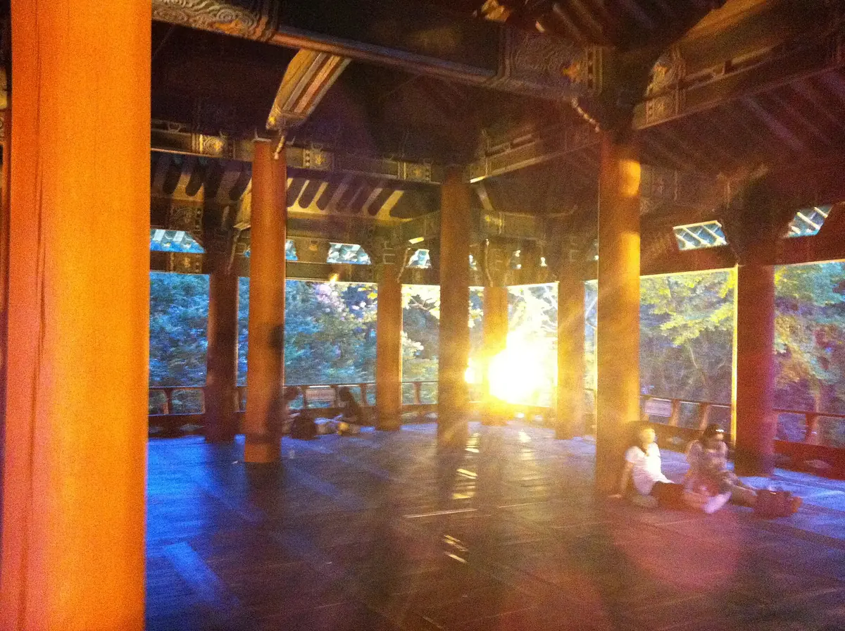 Orange sunset light between the wooden columns of a pavilion, with visitors sitting on the floor looking out
