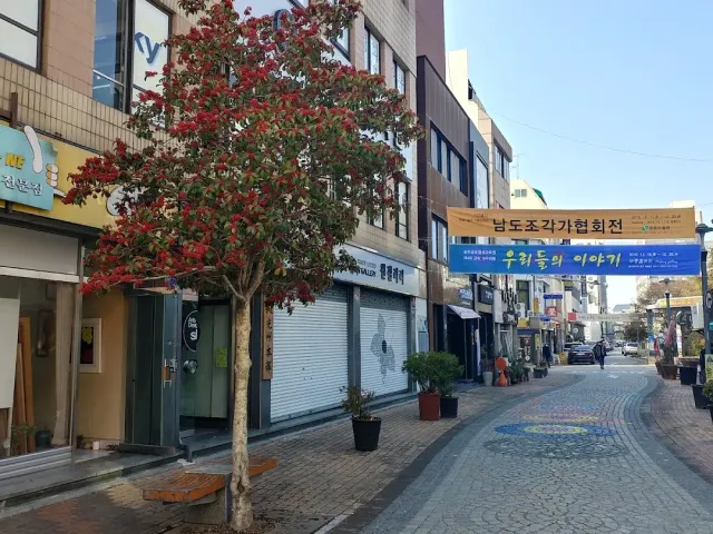 A street of shops with a red leaved tree, a hanging cloth banner and patterned paving