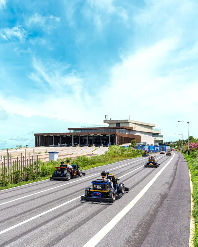 Small racing carts descending a wide asphalt track in front of a low building
