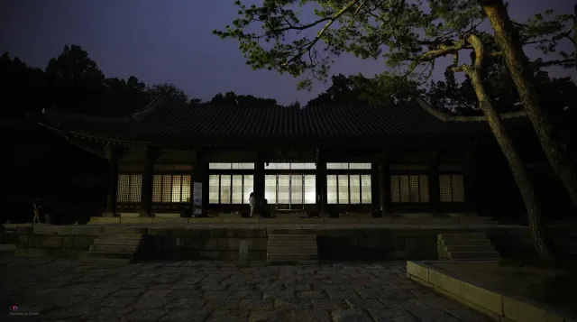 A traditional palace hall lit from within at dusk above a stone courtyard
