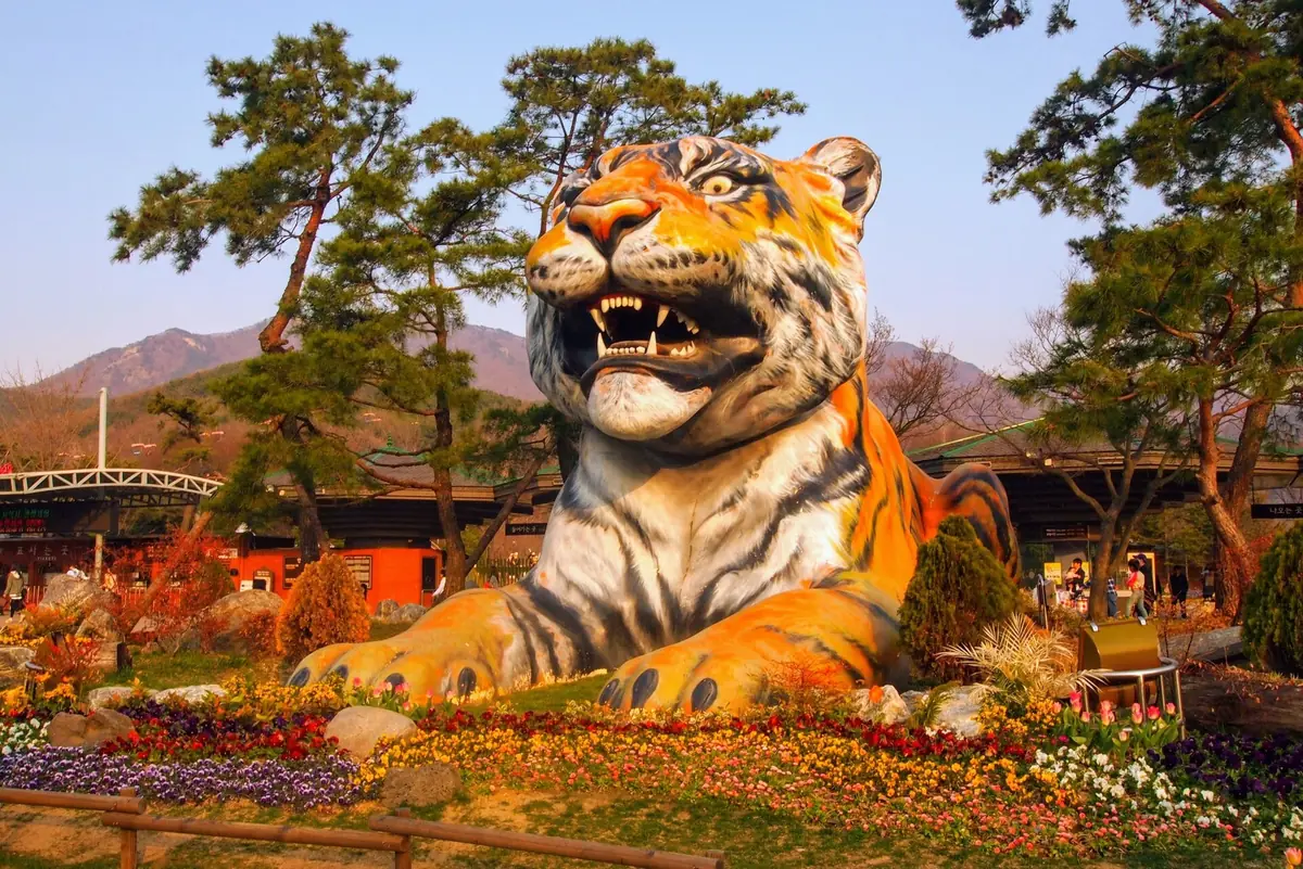 A giant tiger sculpture lying among flower beds, with pine trees and hills behind