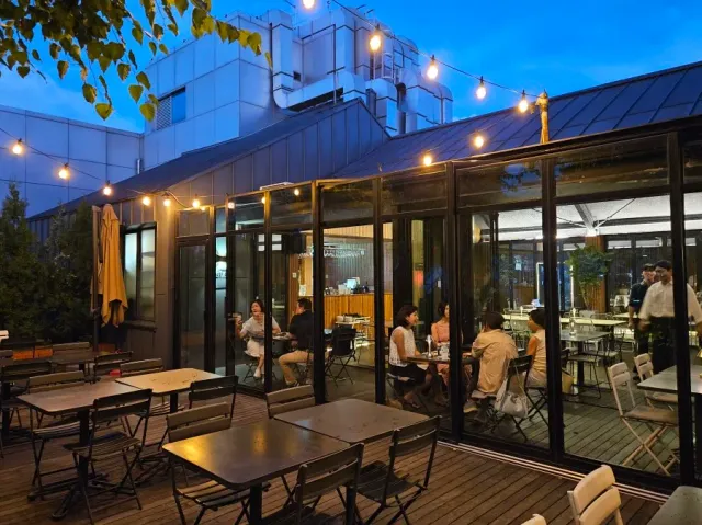 A rooftop terrace in the evening with strings of bulbs, metal tables and guests behind glass