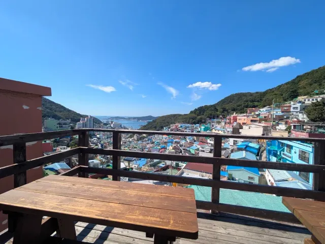 A terrace with a wooden table and railing, facing colourful houses on a slope with the sea behind
