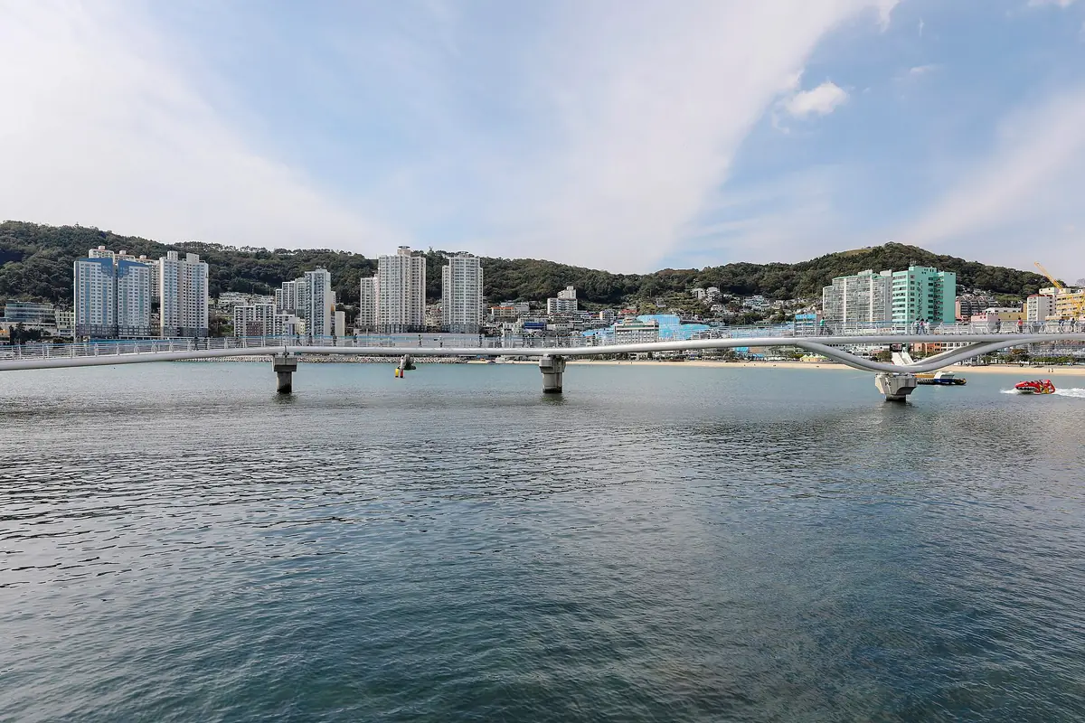 White pedestrian bridge crossing a bay with a wide beach and city towers on a green hillside behind