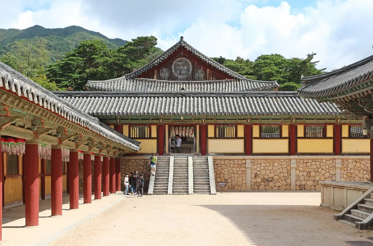 A temple courtyard with a red wooden hall, a stone terrace and stairs, against green hills