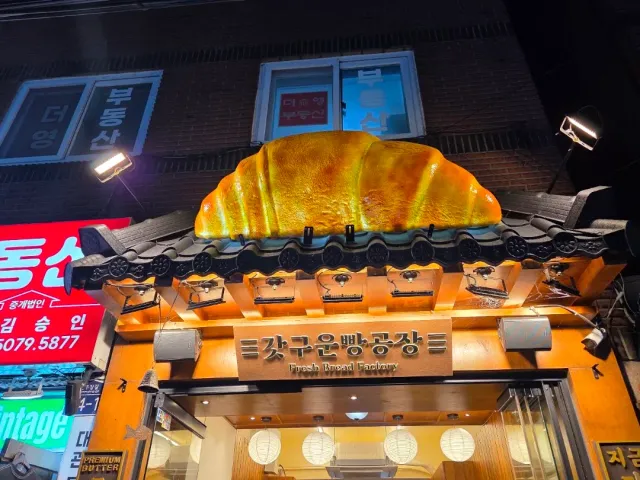An entrance at night with a giant croissant sculpture above a tiled canopy and a lit wooden sign