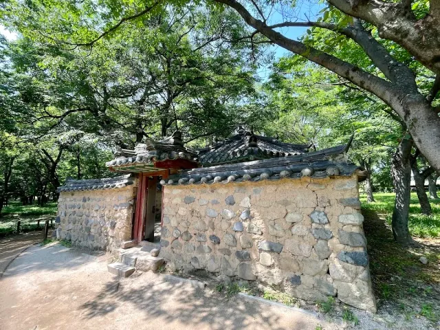 A compound enclosed by a stone wall with a gate and a traditional tiled roof, in the shade of old trees