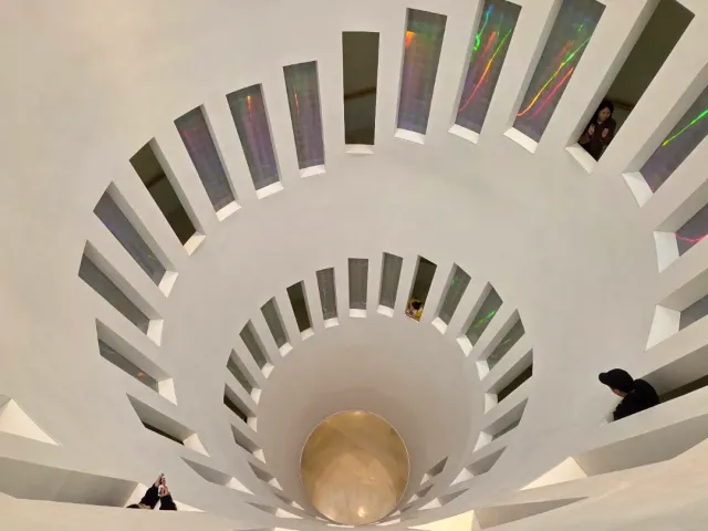 A white spiral opening in the ceiling with narrow rainbow tinted windows and visitors peering in
