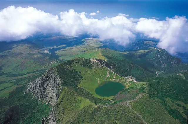 An aerial view of a green mountain crater with a dark lake at its center, clouds around