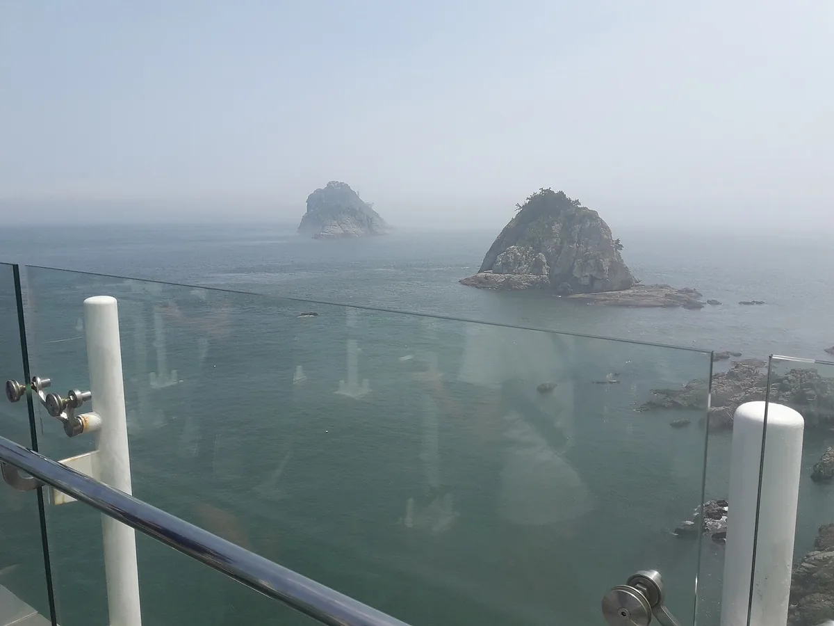 View through the glass rail of a viewing deck towards two green rocky islets in a hazy sea