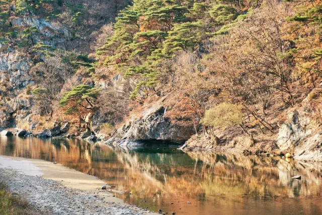 A calm river between rocky slopes and autumn colored trees, a gravel bank in front