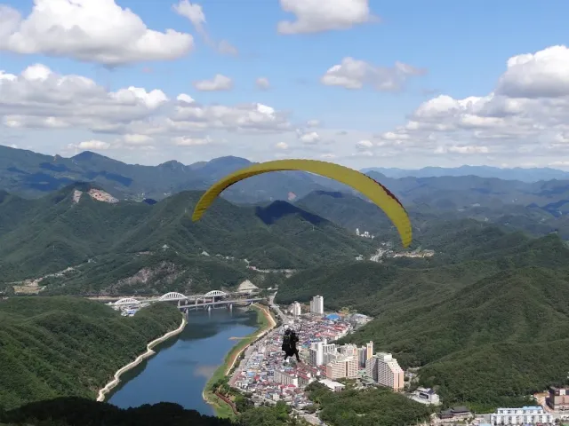 A yellow paraglider in the air above green mountains, a river and a town below